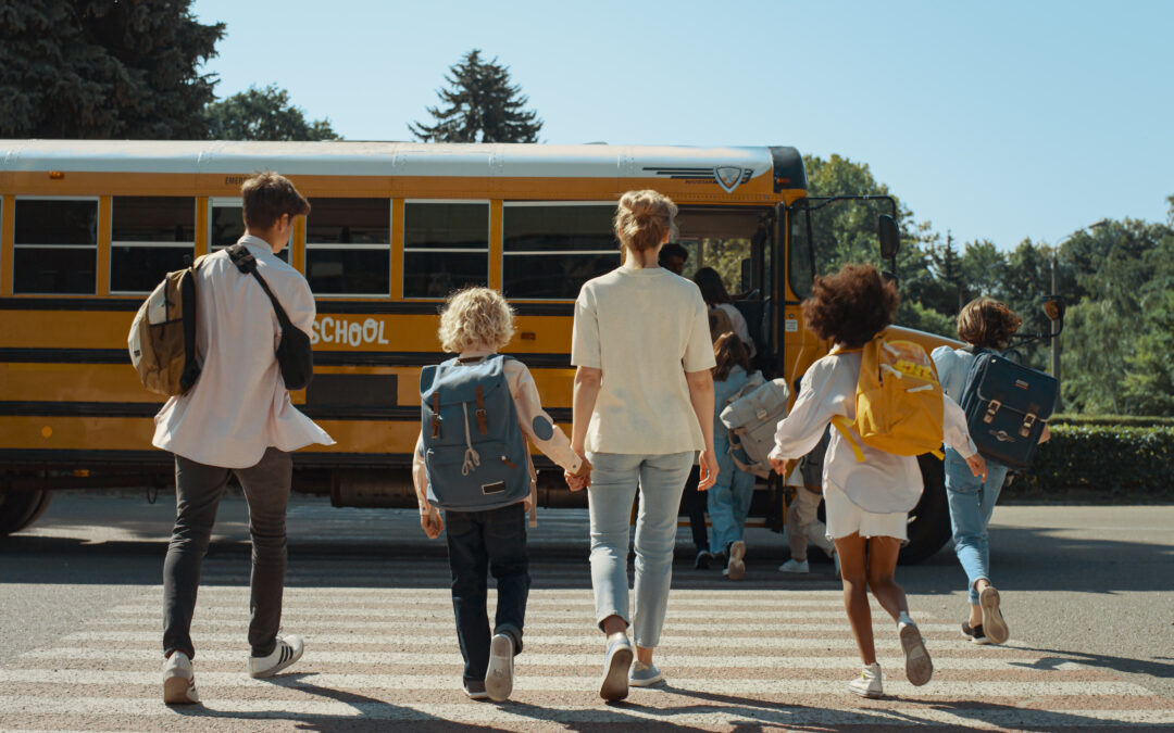 School students walking towards a parked bus to head to school with a mom and her child in the back of the group also walking hand in hand towards the bus.