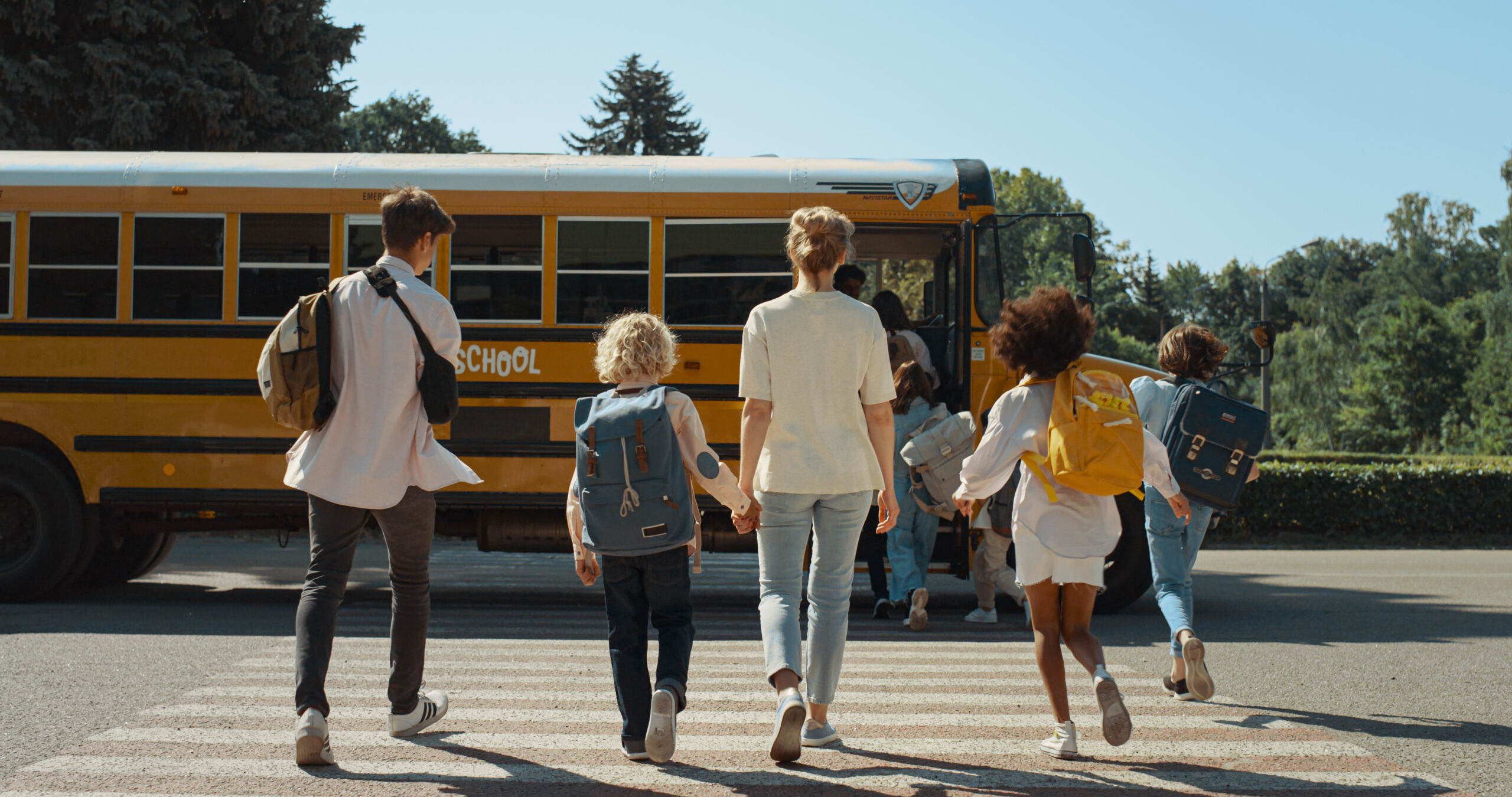 School students walking towards a parked bus to head to school with a mom and her child in the back of the group also walking hand in hand towards the bus.
