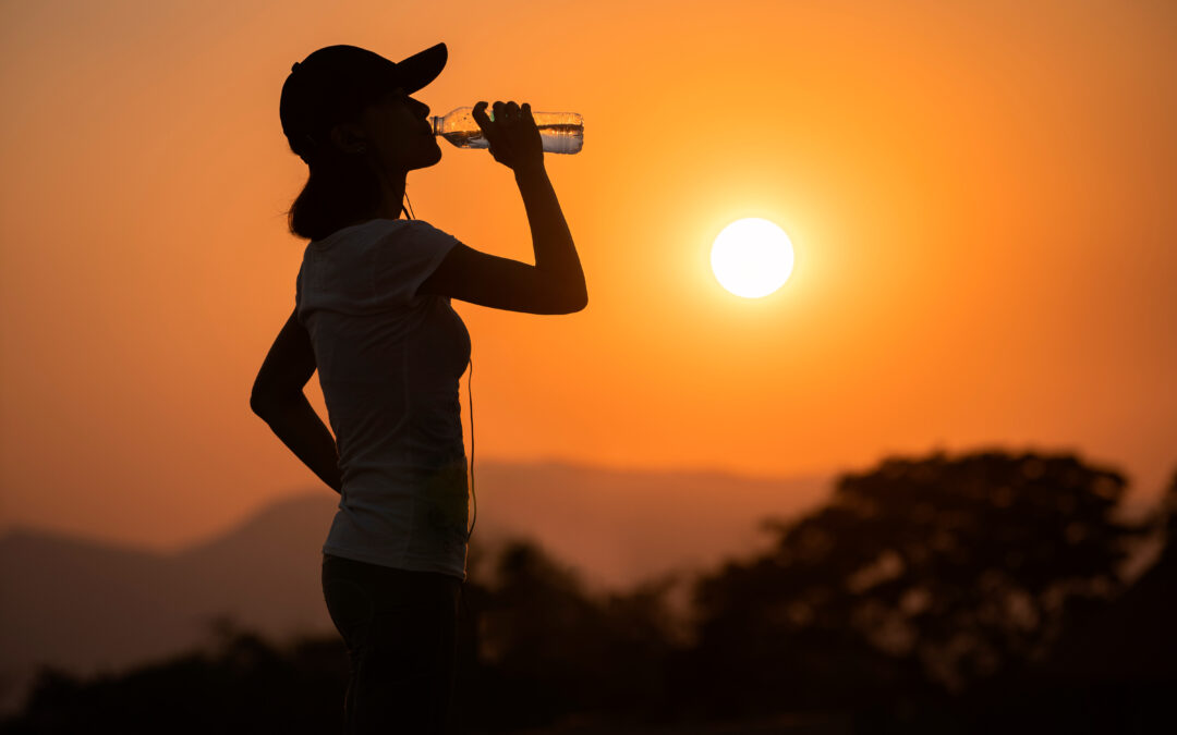A woman wearing a baseball cap stopping to take a sip of water while hiking on a hot, hazy day.