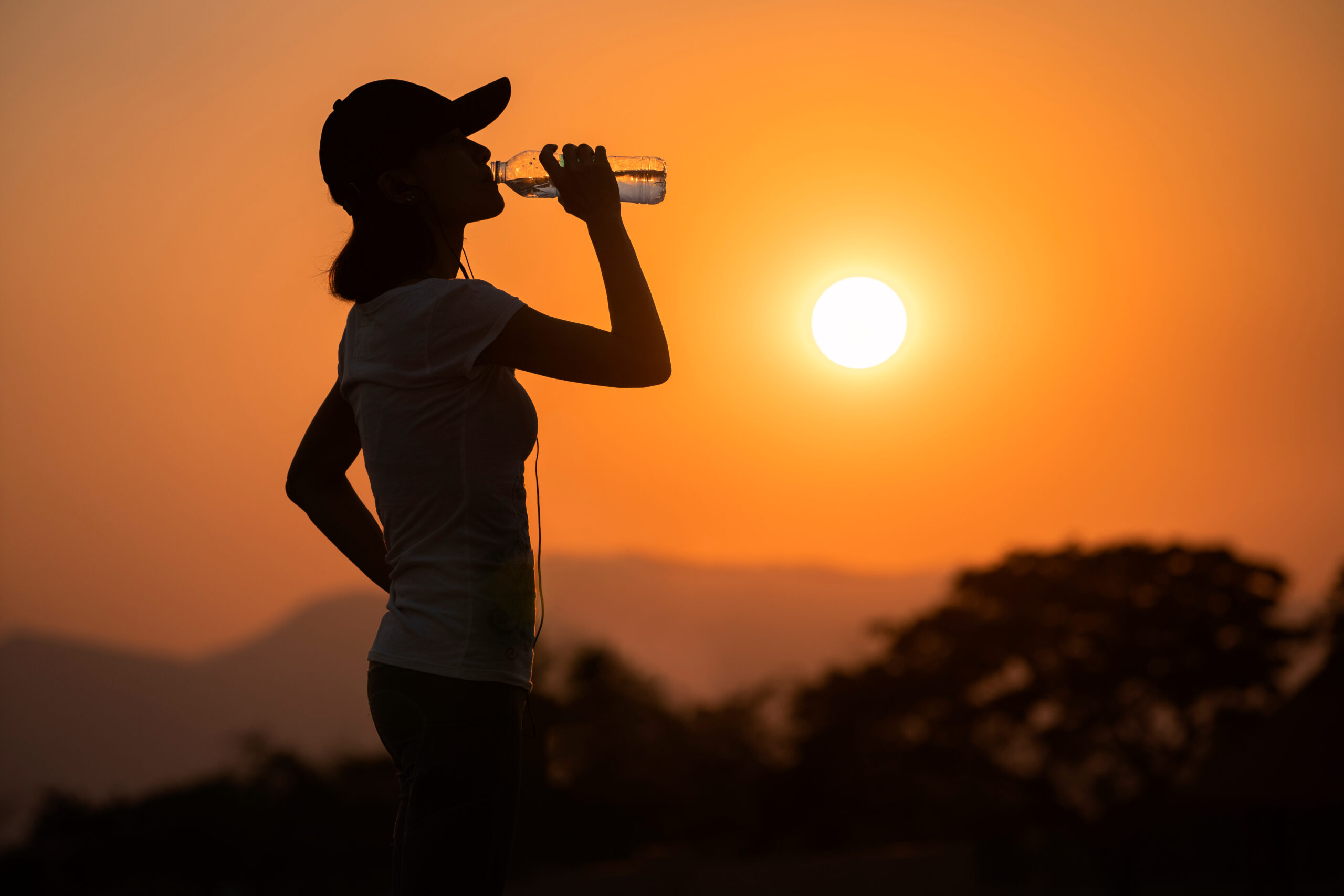 A woman wearing a baseball cap stopping to take a sip of water while hiking on a hot, hazy day.